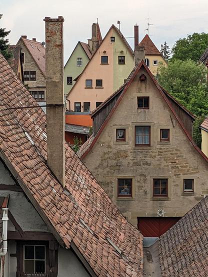 Frontaler Blick auf die steil begrenzten Giebel mehrerer Häuser in einer historischen Altstadt, teilweise mit sichtbarem Fachwerk und Mauersteinen, teilweise mit rosa, grün oder gelb angestrichenem Putz. Dazu das Rot der Dachziegel und etliche Schornsteine.