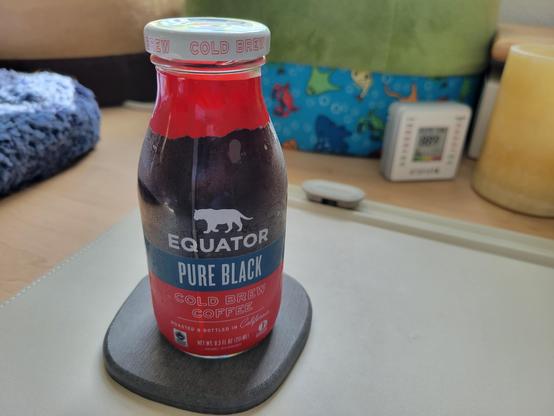 The main focus is a bottle of Equator Pure Black Cold Brew Coffee (8.5 oz) on a gray diatomaceous-earth coaster on an off-white desk mat on a maple desk. In the background are two heated cat beds, a flat pet bed, and an Aranet4 HOME CO2 meter. 

The bottle is clear with condensation. It has a white metal cap and a rough-edged, red border around the neck. The Equator logo appears in white near top-center (jaguar walking, above wordmark Equator in white block letters). 

Text "Pure Black" (white sans-serif block letters) appears below this on a solid-blue band.

The rest of the bottle below the blue band is solid bright red. Text "Cold Brew Coffee" (outline of white sans-serif block letters). A 2-inch thin white line divider separates the next text, "Roasted & Bottled in California" (small white sans-serif block letters, with the state in capitalized script).

Footer text: "Net Wt. 8.5 Fl Oz (251 mL)" (tiny white sans-serif block letters). On the right, tiny wordmark "1% for the Planet" appears, which is a globally recognized certification. On the left, tiny wordmark for "Fair Trade Certified" appears
