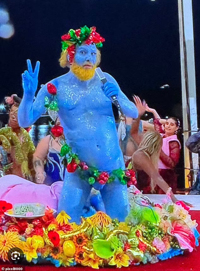 Photo of "naked blue guy" from the 2024 Summer Olympics Opening Ceremonies. A rose garland artfully covers most of his sexy bits. His hair and beard are bushy and bright yellow. He holds a microphone in one hand and flashes a peace sign with the other. He's kneeling on a well-appointed table, and brightly-clothed figures cavort behind him. He is Dionysus, the Greek god of fertility, wine, and pleasure (known to the Romans as Bacchus).