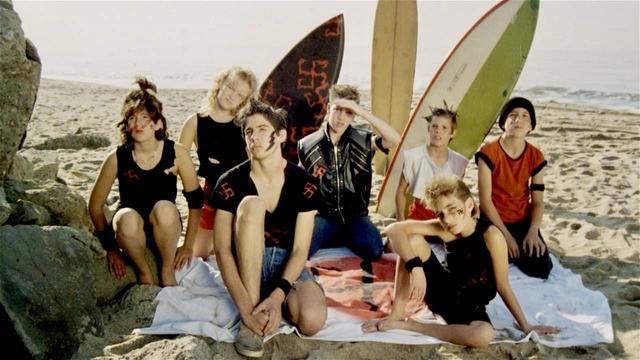 A group of young individuals sitting on the beach in front of surfboards. Some have face paint and wear punk-style clothing.