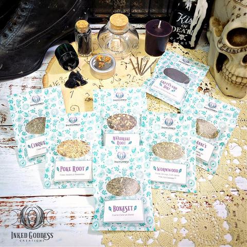 The contents of our exclusive "Dark Witch Kit" product are arranged on a doily-covered tabletop. They include a dark glass jar of black coffin nails; a glass vial of black matches; a clear glass bottle with a cork stopper; a purple "Dark Witch" votive candle; a pouch of graveyard dirt; and 6 herb pouches containing cinquefoil, poke root, mandrake root, boneset, wormwood, and Queen Anne's Lace. This is an exclusive product from Inked Goddess Creations.