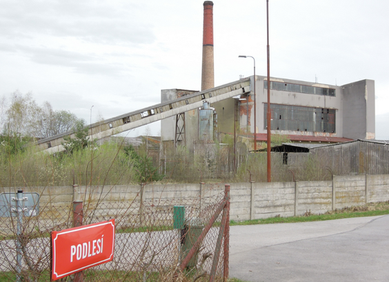 Foto aus dem schlot.at-Archiv (2024) zeigt Fabriksteil der Eisenbahnwerkstätten České Velenic in Tschechien, vermutlich Kraftwerk mit Schornstein und Schrägförderanlage für Kohle, im Vordergrund Maschendrahtzaun und rotes Schild mit Aufschrift "PODLESÍ"