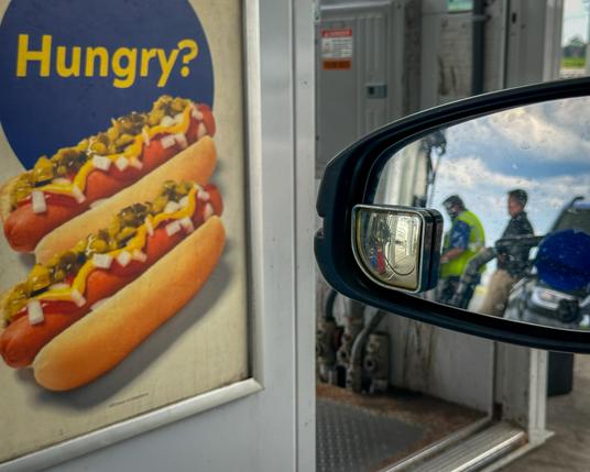 Color photo, composed with two halves in one shot: on the left, a poster with a poster of two hot dogs, garnished to perfection with mustard, relish, and onions. A single word above them taunts you: “Hungry?”

On the right, driver’s side view mirror showing in reflection, the gas pump nozzle in the side of the car and two people, one of whom is the gas station employee, wearing a hi-viz vest.