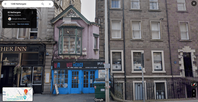Tiny-looking brightly coloured building sandwiched in between two buildings. It has a bay window and pointed roof. To the left is the entrance to a pub and on the right is a block of flats.