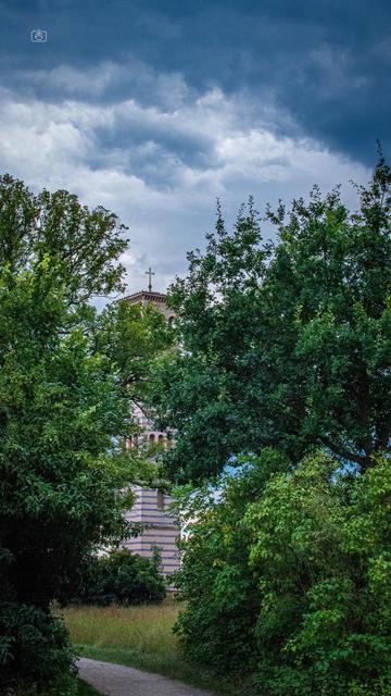 Bell tower of the Church of the Redeemer, partially obscured by tall trees and under a dramatic cloudy sky. Sacrow, Potsdam, 23 Jul 2024. Nikon D5600, Nikkor DX 35 mm ƒ1.8G, ISO 2500, ƒ14, -0.3 ev, 1/250s, polarizing filter