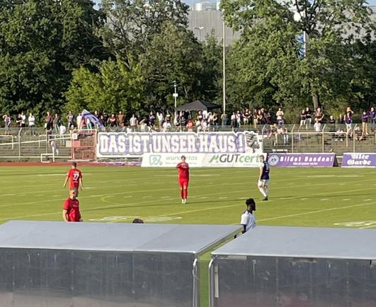 Fußballstadion. Auf einer Bande steht „Das ist unser Haus.“  🎶