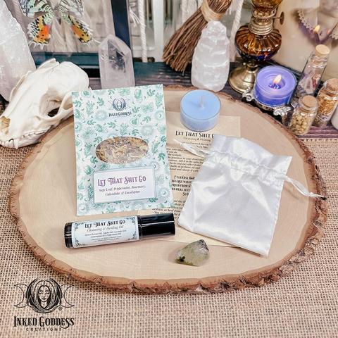 The contents of our exclusive "Let That Shit Go Spell Kit" product are arranged on a raw wood slice. They include a blue Let That Shit Go tealight candle; a pouch of Let That Shit Go herbs, including sage leaf, peppermint, rosemary, calendula, and eucalyptus; a roller ball vial of our exclusive Let That Shit Go Cleansing & Healing Oil; a tumbled prehnite gemstone; kit instructions; and a small white satin drawstring bag. Presented by Inked Goddess Creations.