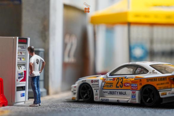 A male stand in front of a cigarette vending machine beside a Nissan S15 Silvia No.23, 2022 Goodwood Festival of Speed by MINI GT in 1:64 scale with 1:64 figure on AREA 164