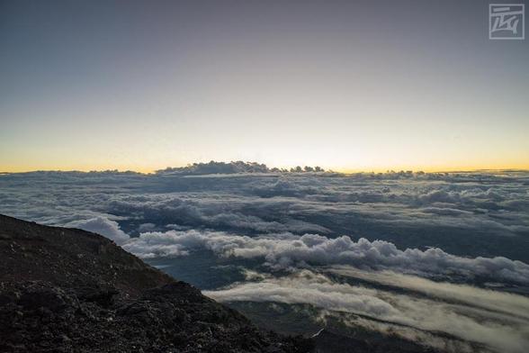 The sun rising above the clouds, seen from the top of Mt Fuji, so there are many clouds below the camera and barren rock in the foreground