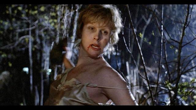 A person with blood on their mouth wearing a torn garment stands in a dark, wooded area.
