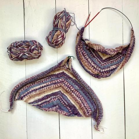 Two knit triangles on circular needles. They're shown on a white plank background, together with 2 balls of variegated yarn in various lilacs.