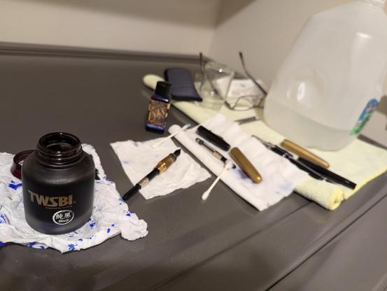 Photograph of three partially assembled fountain pens, bottles of ink, ink-stained paper towels, cotton tips, yellow microfiber cloth, gallon jug of water and other materials used during cleaning and inking spread across the metallic grey top of a clothes washing machine.