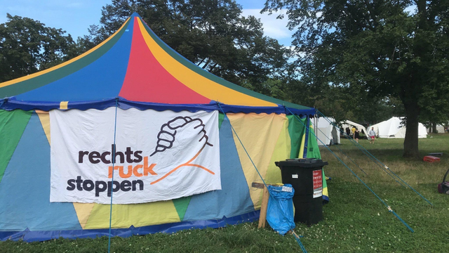 Ein buntes Zelt auf einer Wiese. Das Zelt ist in den Farben Blau, Grün, Gelb und Rot gestreift. An der Vorderseite des Zeltes hängt ein weißes Banner mit der Aufschrift "Rechtsruck stoppen" und einer Illustration von zwei ineinandergreifenden Händen. Im Hintergrund sind weitere Zelte und Bäume zu sehen. Auf der Wiese liegen ein paar blaue Müllsäcke und eine schwarze Mülltonne. Im Hintergrund sind ein paar Personen zu sehen, die zu den anderen Zelten gehen.