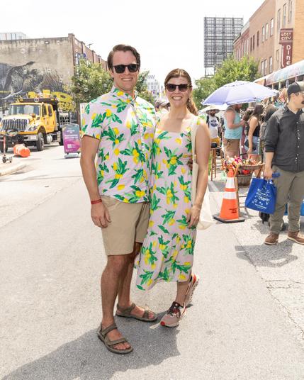 full length street portrait of a smiling man and smiling woman. He is wearing sunglasses, khaki shorts, and a short sleeved shirt with lemons and leaves on it. She is wearing sunglasses and a long sleeveless dress with the same exact lemon and leaf motif. Background is a street fair with pedestrians, parasols, exhibit tents, and buildings.