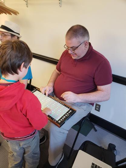 A child and a man play together on a Linnstrument, an electronic instrument with a 2D keyboard. The keyboard has lights on, in various patterns.