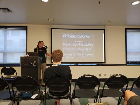 A person is presenting in front of a projector screen, behind a podium. One audience member is visible in the front row. There is a lot of text on the screen.