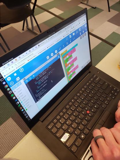 A laptop is visible with a browser open to Music Blocks. Colorful visual code can be seen at right, and a console of text code can be seen at left.