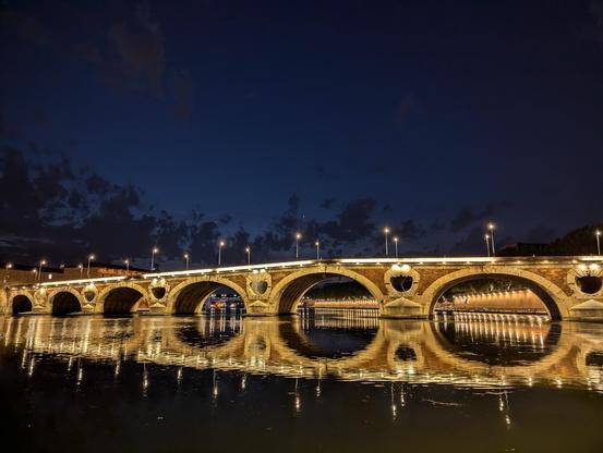 Pont Neuf à Toulouse sur la Garonne pris depuis un quai en contrebas. On voit son reflet dans l'eau sombre de la Garonne. Le ciel est bleu foncé et le pont est illuminé par une lumière chaude, créant un vrai beau contraste.