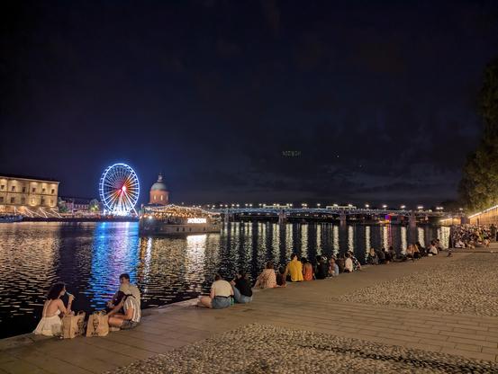 Quai de Tounis et Garonne un soir d'été. Des personnes sont assises le long des quais. Sur l'autre rive on devine l'hôtel dieu et une grande roue, dans le fond le pont St Pierre. Un petit bateau touristique avec des lampions passe sur la Garonne.
