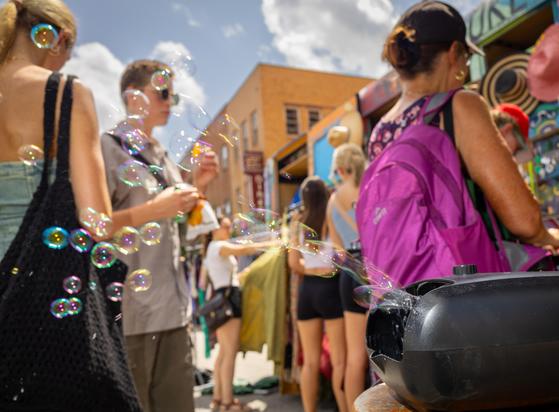 Color photo of a small group of people at a street fair in line to buy food items. In front and to the side of the group is a black soap bubble machine emitting a stream of iridescent soap bubbles past the group. A sunny sky with fluffy clouds is above some building rooflines in the background.
