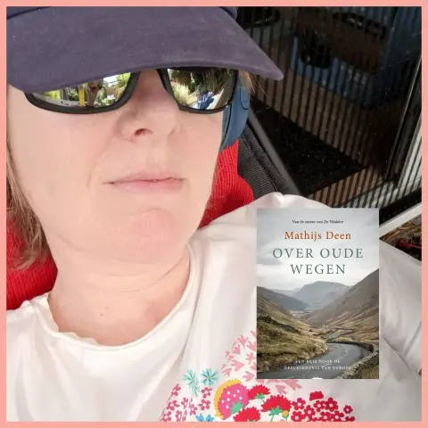 Selfie of white woman with Mirror glasses and blue cap, white tshirt with red pink blue flower print. Lying in reclining chair. 
Insert of the book Over old roads, a history of Europe. Over oude wegen door Mathias Deen 