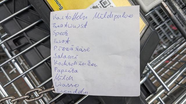 Bildbeschreibung von ChatGPT. 
Das Foto zeigt einen #Einkaufszettel. Der Zettel ist handgeschrieben und enthält die folgenden Artikel:

1. Kartoffeln
2. Bratwurst
3. Speck
4. Brot
5. Pizza-Käse
6. Salami
7. Kochschinken
8. Paprika
9. Milch
10. Wasser
11. Windeln

Der Einkaufszettel ist auf einem weißen Blatt Papier geschrieben und liegt auf dem Griffbereich eines Einkaufswagens. Man kann auch Teile des Einkaufswagens sehen, einschließlich der Kette, die zum Verriegeln der Wagen verwendet wird. Der Hintergrund zeigt die typische Metallstruktur eines Einkaufswagens.