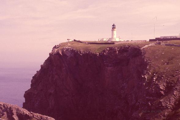 Looking across at a cliff top with a lighthouse, a fog horn to its left, and some low buildings. The cliff top is grassy, but with sudden crags. The sea can be faintly seen in the haze.
