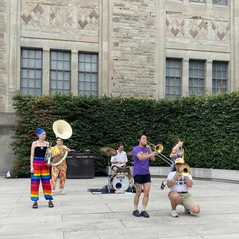 Trash Panda Brass playing outside the Royal Ontario Museum. Two mellophone players, two trombonists, a sousaphone player and a drummer, all brightly coloured and lively as they play on a sunny August afternoon. 