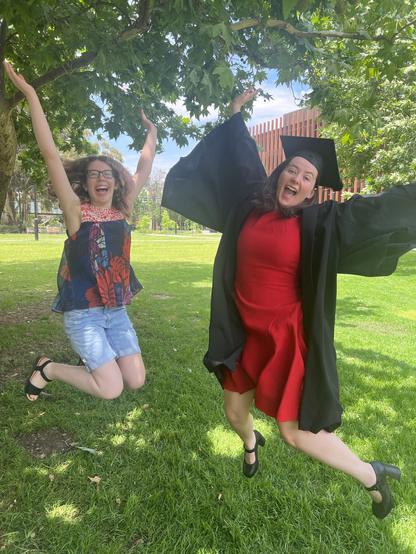 Two women jumping in the air. One has a university graduation cap and gown on.