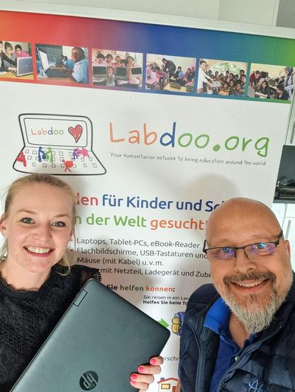 Das Foto zeigt 2 Personen vor einem Ausschnitt eines Labdoo Rollups. Links steht Leandra vom Labdoo Hub Hamburg. Sie hält einen zugeklappten Laptop in der Hand. Rechts steht Phillip von der spenden Firma HPM aus Hamburg. Beide schauen in die Kamera und lächeln.