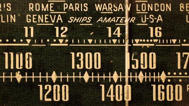Old fashioned radio tuning band, names of various European cities, frequency numbers,  Black background with light coloured names and numbers 