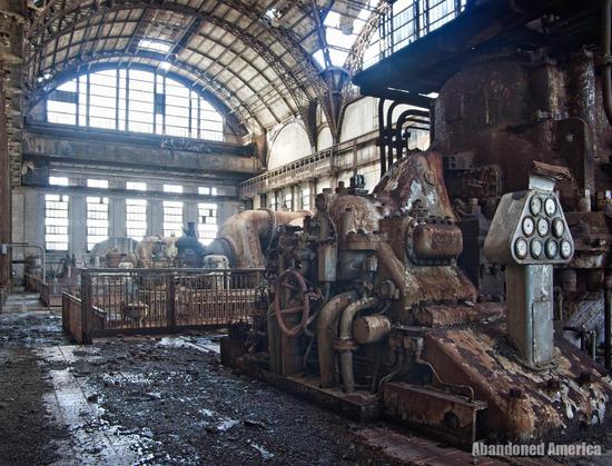 A massive turbine hall atrium with large chunks of concrete missing from the ceiling. A large, rusty turbine is in the foreground. 