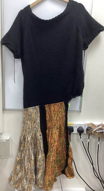 A long view of a dress. Half the skirt is completed. The dress is a black top and the skirt has two coloured sections. The first is white, blue, gold and yellow. The second colour section is red, green, orange and yellow. There is a black band between the two coloured sections.