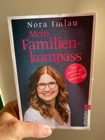 The foto shows a hand holding a book. On the book cover is the title „Mein Familienkompass“ and the name of the author Nora Imlau. It also shows a picture of a smiling women with long hair and glasses.