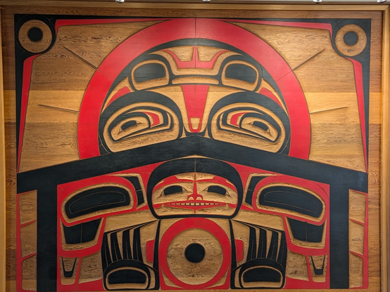A large wall panel carved and painted in the black and red ovoid style of the First Peoples in the Pacific Northwest shows a tutelary or protective entity or spirit, whose countenance comprises representations of salmon, guarding or observing a house in which a humanoid or ursine person faces the viewer with paws or hands raised and with a kind of wistful smile.