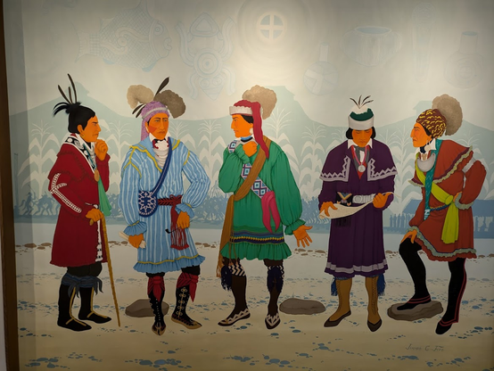An oil-on-canvas painting shows five life-size, relative naturalistic representations of standing tribal representatives in distinct, festive, formal-looking clothing, apparently in deep deliberation and conversation. One of them studies what's written on a large paper, which is maybe what they're dealing with. Superimposed on the mountains in the background one sees shapes of corn, and in front of these are silhouettes of people fighting, a longhouse, other people seemingly waiting. In the pale sky, you can barely see celestial symbols, a fish, a frog, and several pitcher-like items. 