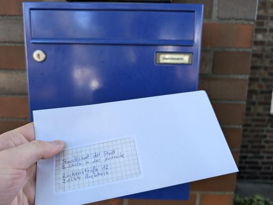 Hand holding a letter addressed to the Standesamt (civil registration office) of Buchholz. Out of focus in the background is a blue mailbox with the text “Standesamt”.