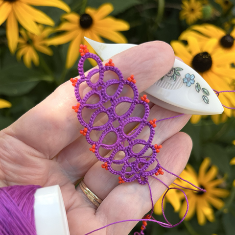 A purple handmade lace in tatting with bright orange beads, held on a hand outside near yellow Rudbeckia hirta (black-eyed Susan) flowers. A white tatting shuttle is also in the hand with tiny flower stickers on it, and the ball of thread.