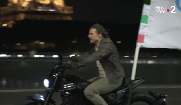 Photo de Tom Cruise conduisant une moto avec à l'arrière, le drapeau des JO de Paris. On peut voir un bout de la tour Eiffel en fond dans la nuit.