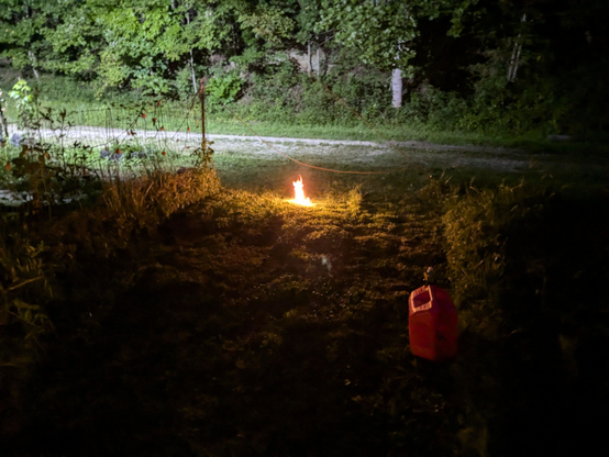 A dark patch of grass with flames leaping out of a hole in the ground.  A red gasoline jug can be seen nearby.