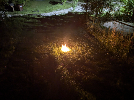 A dark patch of grass with flames leaping out of a hole in the ground.