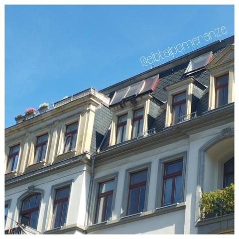 Die zwei obersten Etage eines Gründerzeithauses. Von der Dachterrasse sieht man nur die Blumenkästen am Geländer, deren rosafarbene und weiße Blumen vor dem makellos blauen Himmel leuchten.