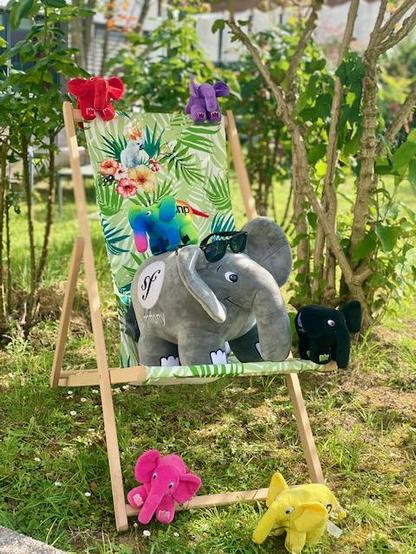 A large gray Symfony elePHPant in a garden with a pair of sunglasses on his head, sitting on a deck chair with a tropical print and surrounded by small elePHPants of many colors.
