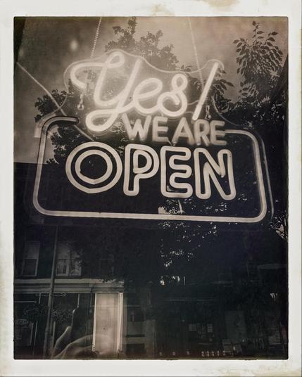 Neon sign in a shop window saying “Yes! WE ARE OPEN. A vintage black and white filter has been applied.