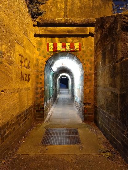 The photo shows the pedestrian railway underpass in Nuremberg Reichelsdorf from the east. The exit area consists of a plastered wall on the left side, which is sprayed with graffiti. On the right, a partially unplastered sandstone wall can be seen. The arched passageway directly below the railway tracks is only 1.1m wide and 2.5m high in the middle. The brick walls of the passageway are also sprayed with graffiti. The exit area is illuminated by a sodium vapour lamp, whose yellow light forms a strong contrast to the cold lighting with fluorescent tubes in the passageway
-
Auf dem Foto ist die Fußgänger-Bahnunterführung in Nürnberg Reichelsdorf in östlicher Blickrichtung zu sehen. Der Ausgangsbereich besteht auf der linken Seite aus einer verputzten Mauer die mit Graffiti besprüht ist. Rechts ist eine teilweise unverputzte Sandsteinmauer zu sehen. Der bogenförmige Durchgang direkt unterhalb der Bahngleise ist nur 1,1m breit und in der Mitte 2,5m hoch. Die gemauerten Ziegelwände des Durchgangs sind ebenfalls mit Graffiti besprüht. Beleuchtet wird der Ausgangsbereich mit einer Natriumdampflampe, deren gelbes Licht einen starken Kontrast zu der kaltweisen Beleuchtung mit Leuchtstoffröhren im Durchgang bildet