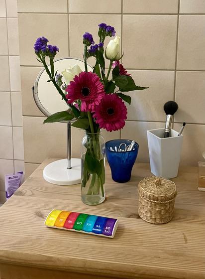 Ein Foto aus meinem Badezimmer. Auf der Kommode steht vorne eine Pillendose in Regenbogenfarben. Dahinter steht ein Strauß aus pinken Gerbera, weißen Rosen und lila Irgendwas.