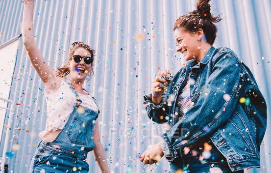 Two women, one in overalls, and one in jean jacket, outside, laughin, dancing and throwing confetti/sprinkles in the air