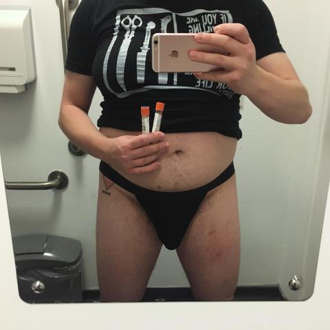 A trans man wearing a black jock strap takes a photo of himself in the mirror of a toilet in a sexual health clinic. He has a tattoo on his left hip and his black t-shirt is pushed up to show the hair on his belly. In one hand he's holding his phone, in the other he's holding the tubes containing his throat and rectal swabs.