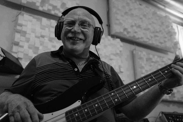 A cool looking bloke in his late 60s with a white moustache and glasses. He has headphones on and is playing an electric bass guitar.