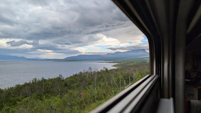 View out of the train window onto the grey Lake Torneträsk. High mountains and clouds in the background. Small trees on the shore in the foreground.